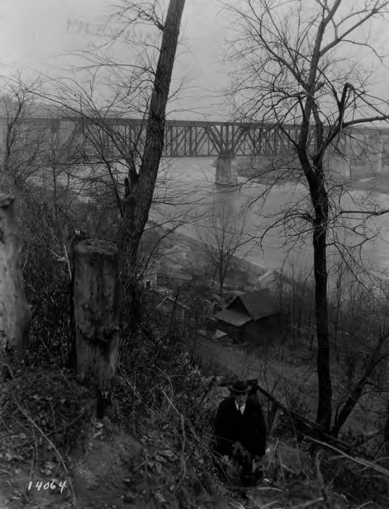 A man in a suit is walking toward the camera up from the steep bluff along the Mississippi River. At the bottom of the bluff is a wooden house and in the distance is the original Washington Ave bridge.