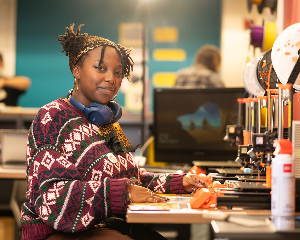 A student in a patterned maroon sweater sits at a desk in front of a 3D printer