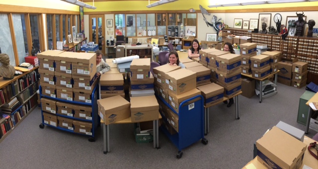 A panoramic photo showing four staff people dwarfed by the 80 boxes of the recently processed Outfront MN collection.