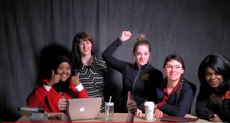 Group of students poses in front of the backdrop of the 1:Button Studio. 5 are sitting around a table. One person giving two thumbs up is out of frame; only their hands can be seen.