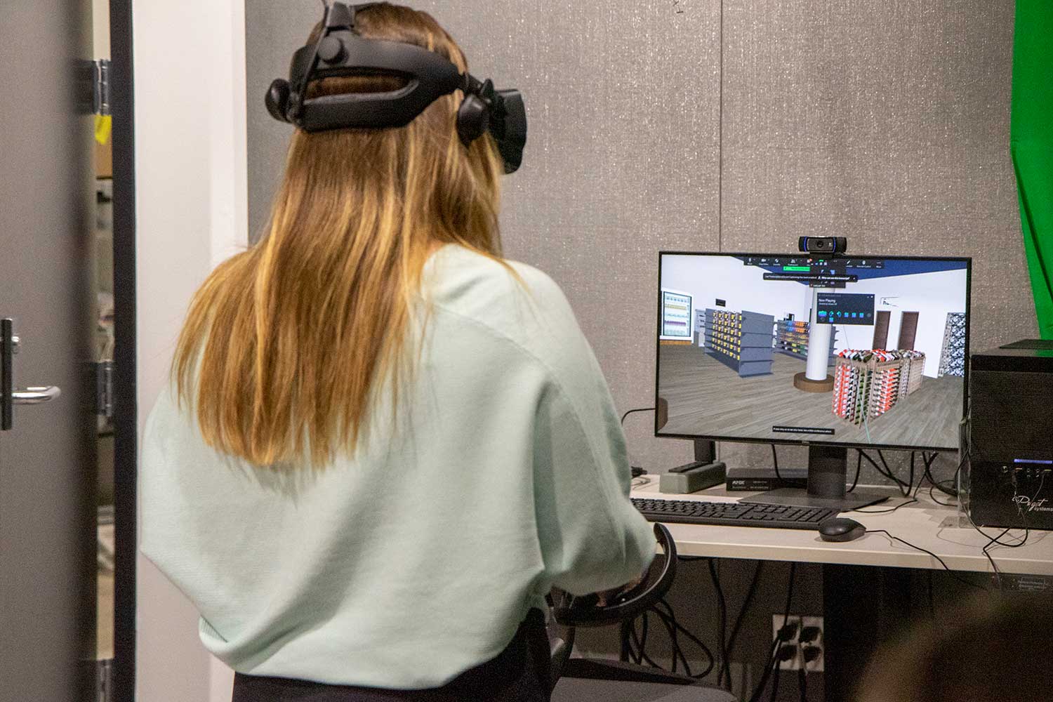 Student wearing VR headset in front of computer, which displays a simulated retail space