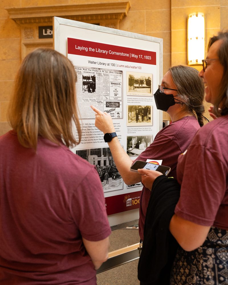 Three staff members in Walter at 100 T-shirts gather in front of a poster that reads, "Laying the Library Cornerstone"