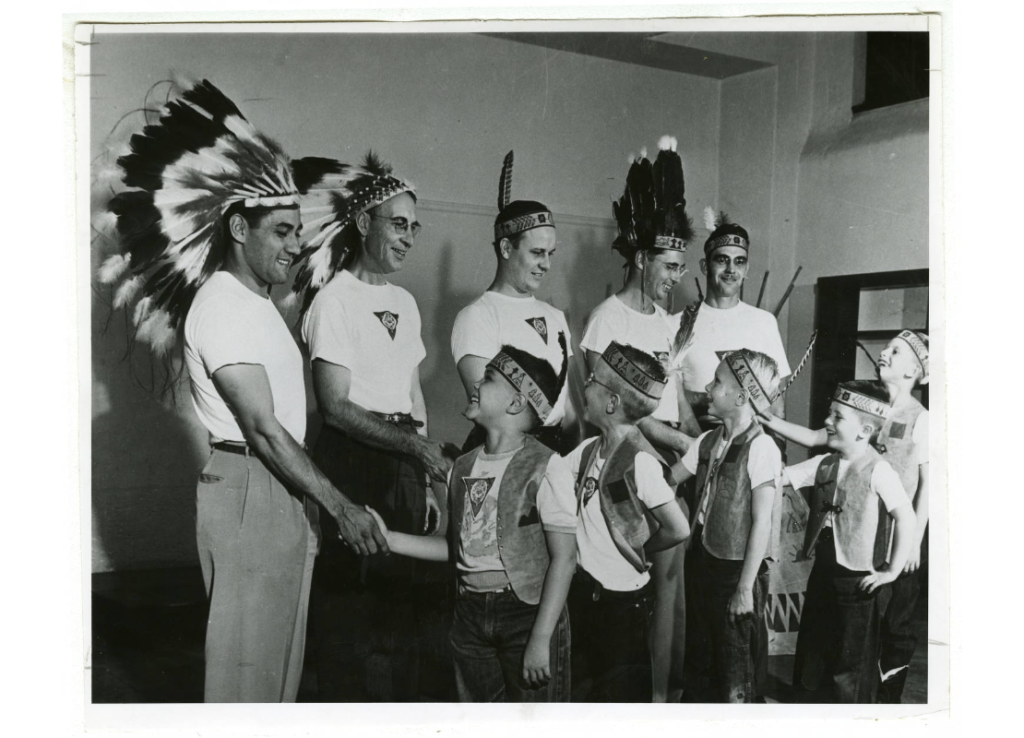 Men and boys wearing Indian headbands and feathers shaking hands, created between 1940 and 1969.