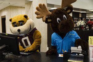 Goldy Gopher mascot typing at a computer next to a moose mascot holding a phone at the front desk of Wilson Library