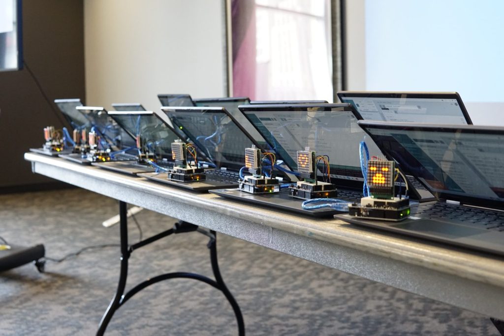 A line of laptops on a folding table. LED matrices showing different patterns are lined up at the front of the table.