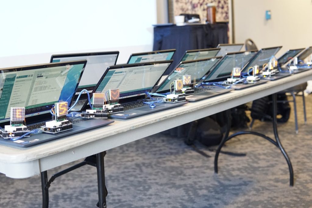 A line of laptops on a folding table. LED matrices showing different patterns are lined up at the front of the table.