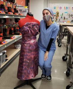 A student in scrubs stands next to a dress on a dress form. The dress pattern is made from magnified photos of skin cells.
