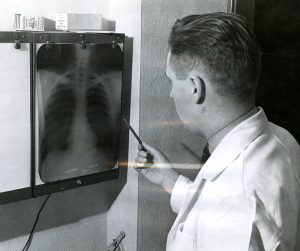 A white man in a white lab coat looks at an x-ray of a chest while pointing at the heart.