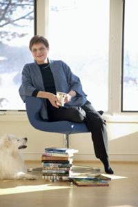 The author seated in a bright room with her fluffy white dog and a stack of her books on the floor beside her