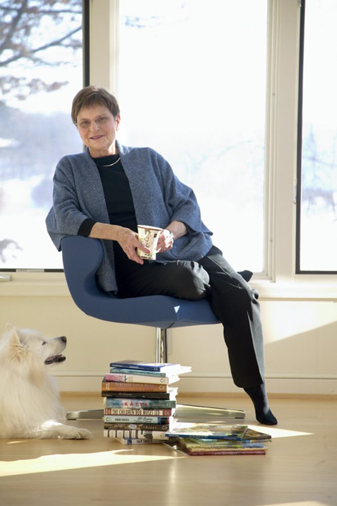 The author seated in a bright room with a stack of her books on the floor beside her