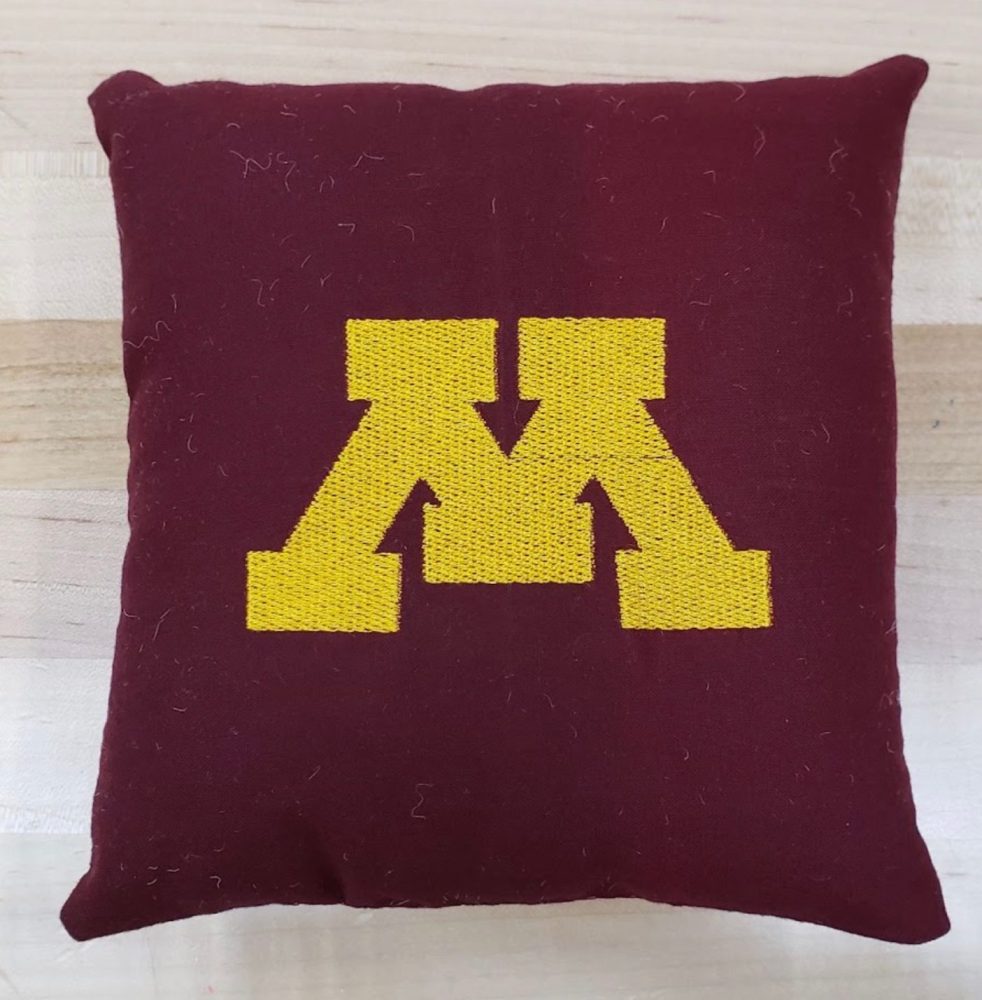 A small maroon pillow with the yellow Block M logo embroidered on it.