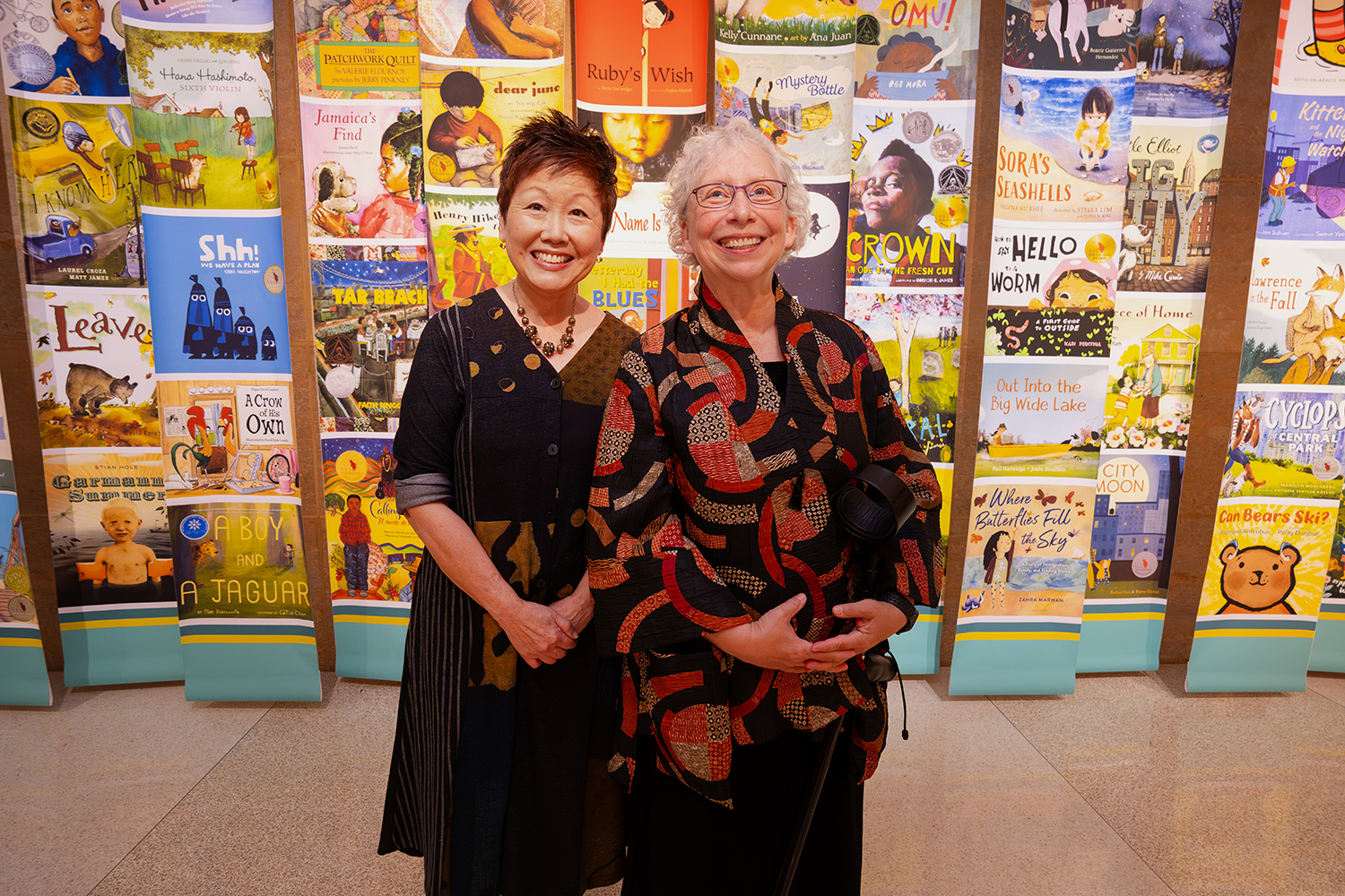 Two women stand side by side in front of a wall covered in images of colorful children's book covers at Elmer L. Andersen Library