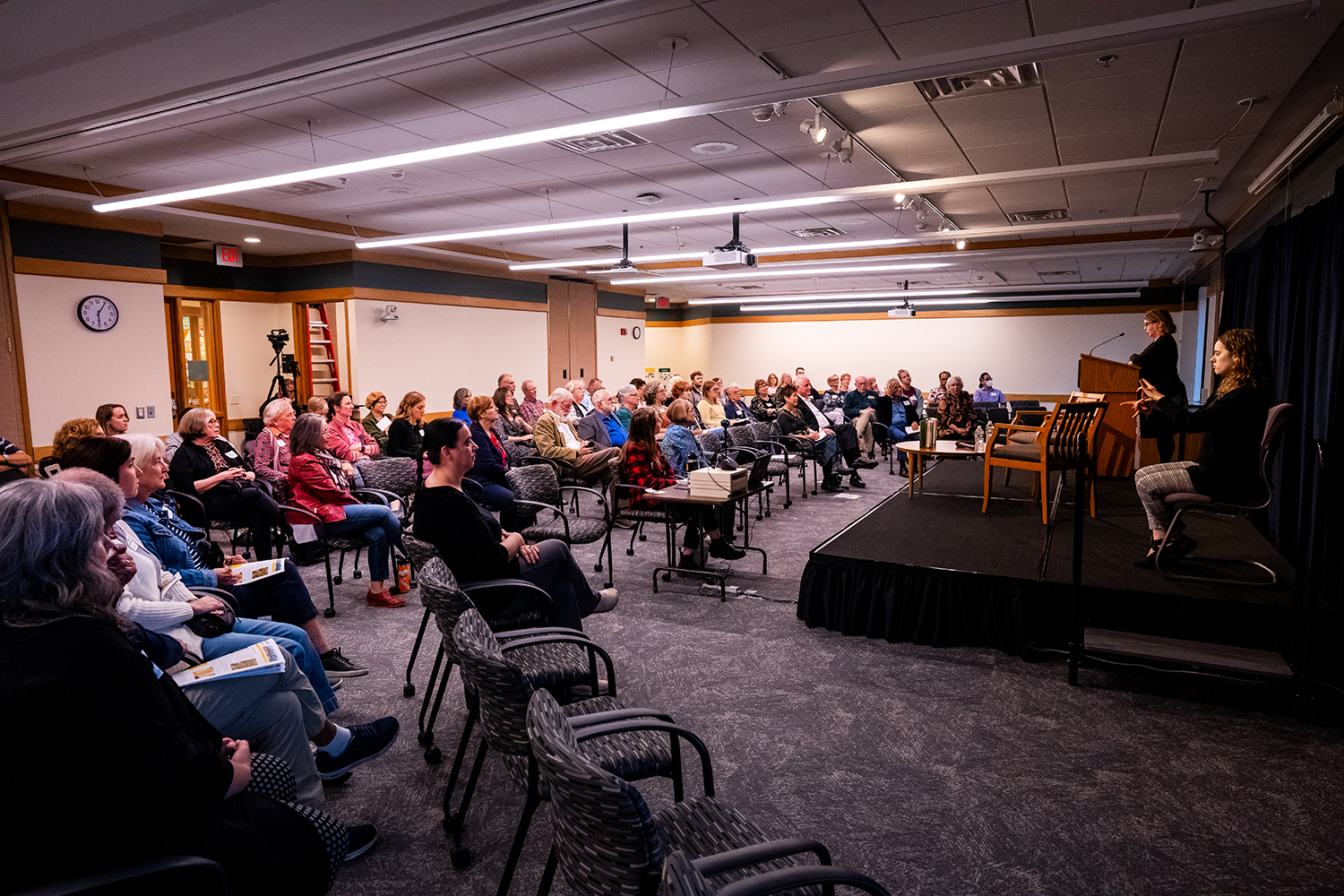 Room full of people seated in chairs in rows facing a stage where Dean Lisa German addresses the audience at a podium