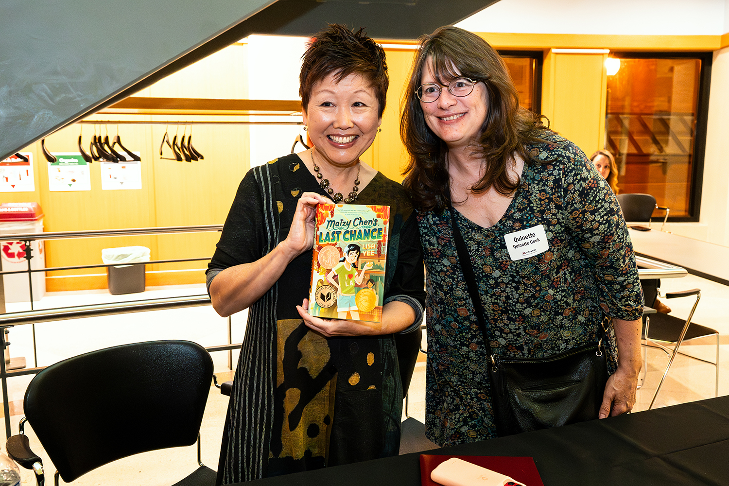 Author Lisa Yee holds a copy of her book Maizy Chen's Last Chance while an attendee stands next to her. Both smile at the camera