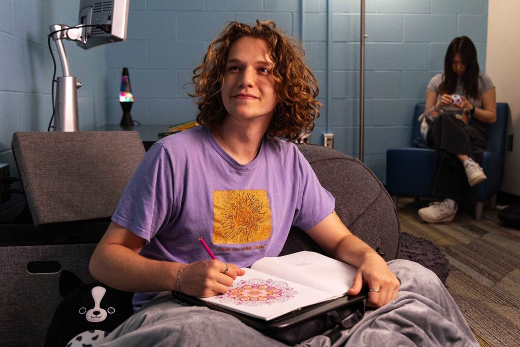 A student relaxes in a wellness room in the library with a blanket and coloring book on their lap. The room is lit with soft lighting