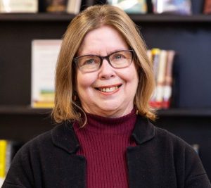 Headshot of woman with shoulder length hair and eyeglasses wearing a turtleneck and jacket in front of a book display in Wilson Library