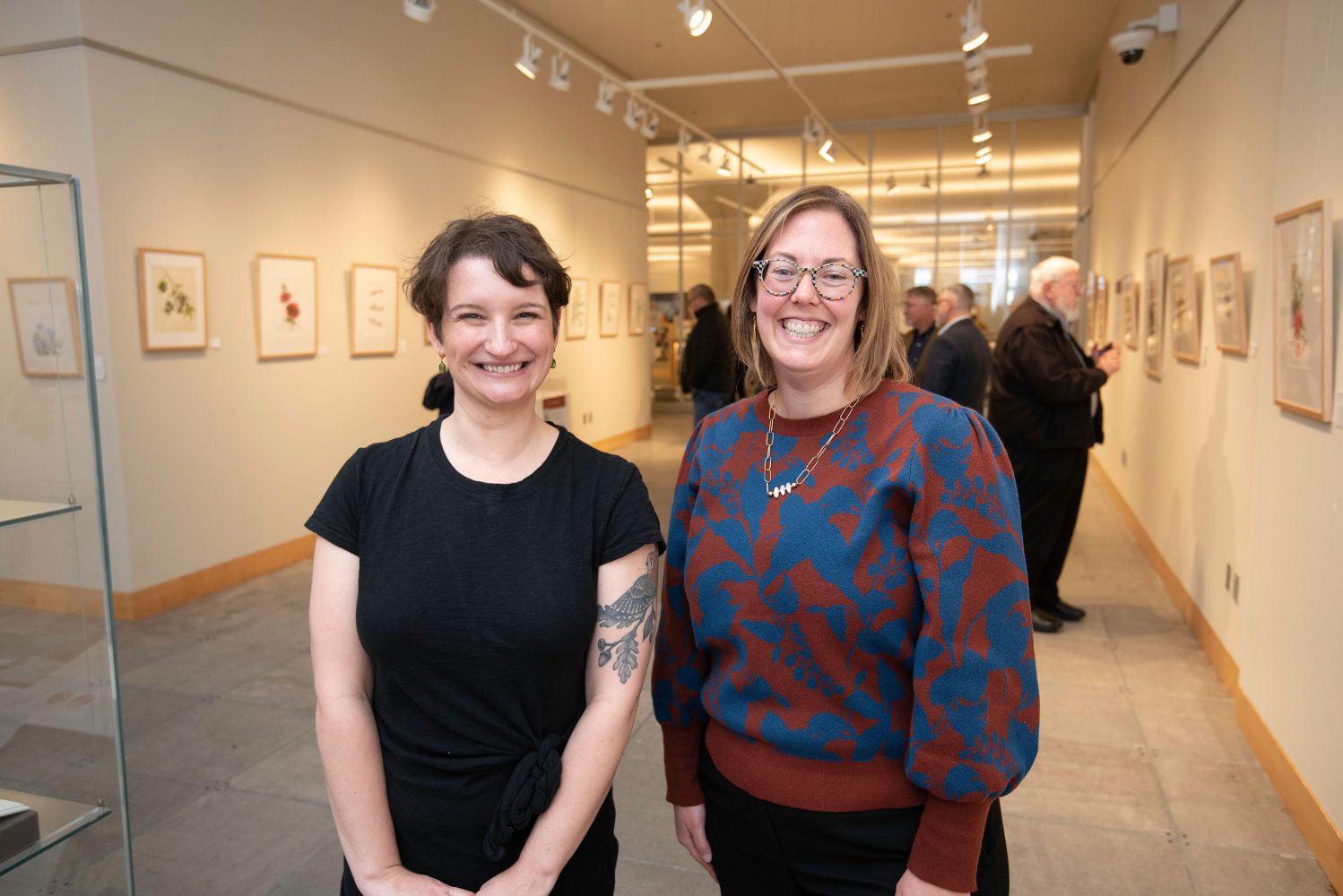 Two women stand smiling side by side in the exhibit space for Flora and Fauna illustrata at the Minneapolis Central Library