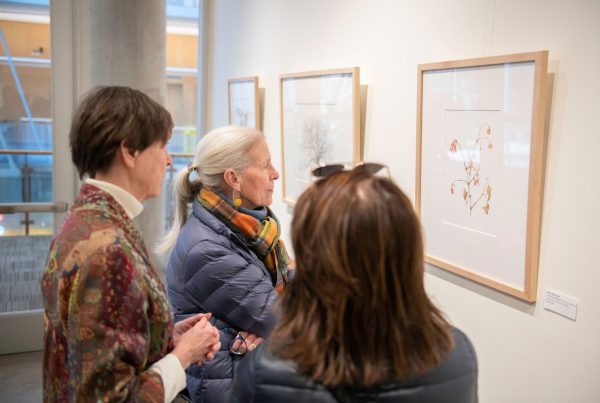 Three women stand in front of a framed drawing of a plant and study it carefully