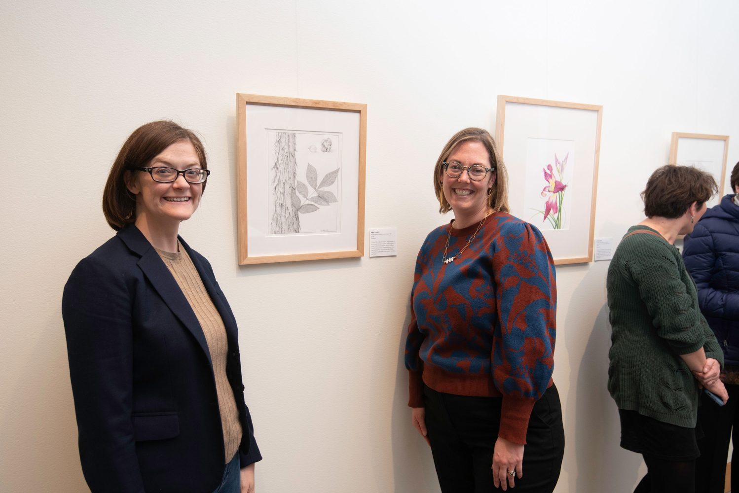 Two women with short hair and glasses stand and smile in front of a framed sketch of a botanical artwork hung on a library gallery wall