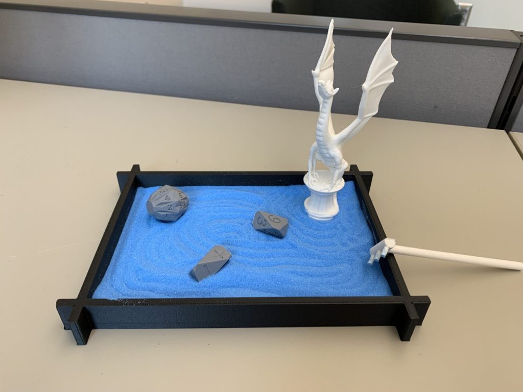 A 3d printed sand garden with blue sand. In the garden are three rock-shaped dice and a white dragon statuette.