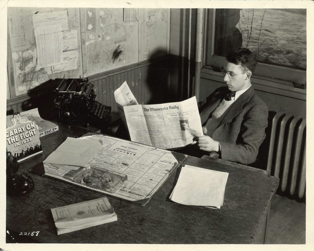 Man sitting at a desk wearing glasses and holding a copy of the Minnesota Daily open in front of him