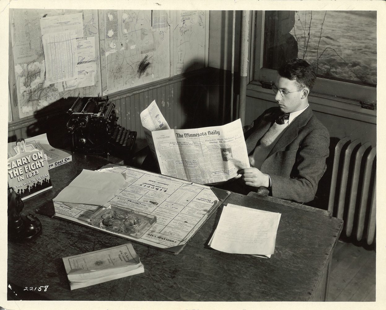 Man sitting at a desk wearing glasses and holding a copy of the Minnesota Daily open in front of him