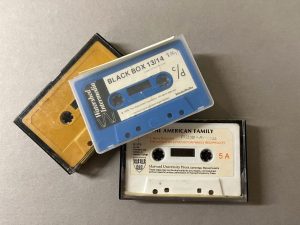 Three audio cassette tapes piled up, one is blank yellow, one is blue with the title “Black Box,” and one is white with the title “The American Family.”