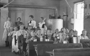 Black and white photograph of young students sitting at school desks, eating out of cups. Four young adult women stand in the background of the classroom