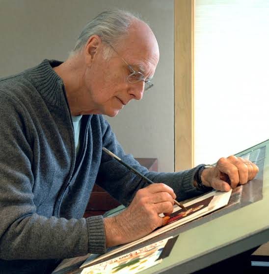 Profile of man sitting in front of a slanted desk where he is looking down and illustrating or painting a picture