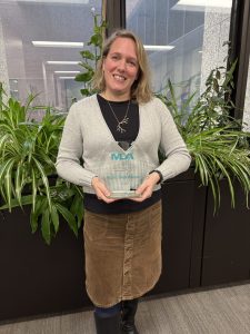 Nicole Theis-Mahon, a white woman wearing a black shirt, grey cardigan and brown skirt, Evidence Synthesis Librarian and HSL Collections Coordinator, holds her plaque honoring her as the 2025 Jean Sayre Innovation Award recipient from Midwest Chapter of the Medical Library Association.