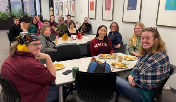 A room with around 20 people sitting and smiling at the camera at a potluck with multiple tables.