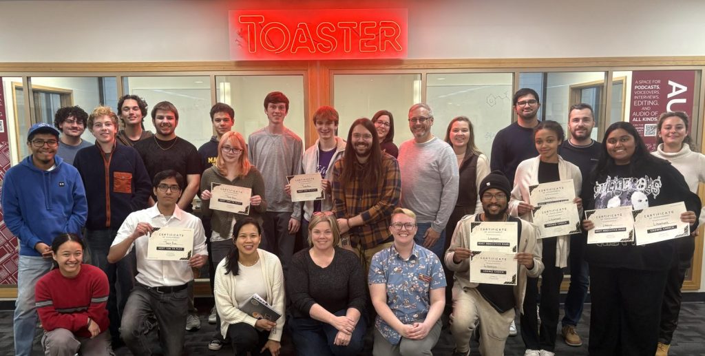 Students and judges for the Tinker @ The Toaster Game Jam challenge gathered under the Toaster neon sign for a group photo.